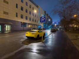 A vibrant yellow sport car is conspicuously parked on a wet city street at dusk, reflecting the glowing streetlights and the illuminated facade of a modern building. The low-angle shot captures the photo