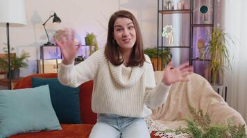 Pretty woman smiling friendly at camera, waving hands gesturing hello, hi, greeting at home on sofa video