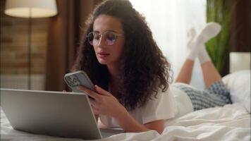 Woman in bedroom using laptop relax on bed Caucasian girl call mobile phone online shopping order delivery internet purchase at home smiling attractive female talk cellphone calling friend communicate video