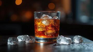 A glass of amber-hued beverage, with ice cubes, stands on a dark surface. photo