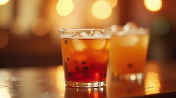Two elegant glasses filled with a refreshing beverage, garnished with ice cubes and small dark berries, stand on a polished table against a softly blurred background illuminated by warm photo