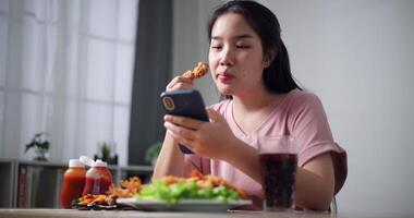 Relaxed young woman enjoys crispy fried food while casually scrolling through mobile phone in living room at home. video