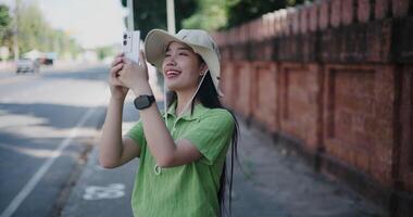 Mano disparo, joven asiático mujer en verde camisa y sombrero en pie en el lado de el calle disfrutando el ciudad ver y tomando fotos con móvil teléfono. video