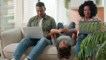 Addicted family use gadgets devices little kid child son boy lying upside down on sofa play game on mobile phone smiling African American parents working laptops different gen using modern tech video
