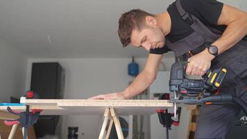 Worker in workwear using handheld cordless electric jigsaw to cut wooden board in garage. Skilled carpenter working in workshop. Man doing DIY home renovation video