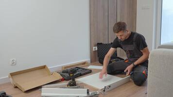 Man in workwear assembles cabinet drawers using tools on living room floor. Concept of DIY home improvement and furniture installation video