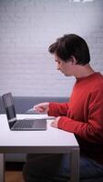 vertical Young man sitting at a white table, using a laptop to book flight tickets online while playing with a toy airplane, blending leisure and travel planning in a comfortable home office setting video