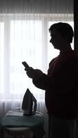 vertical Silhouetted man using a smartphone while ironing clothes by a window at home, blending household chores with modern mobile technology in a comfortable indoor setting video