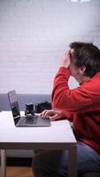 vertical Photographer experiencing burnout and frustration while working on laptop, covering face with hand, with camera and lens on desk suggesting creative block and work stress video