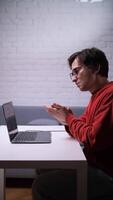A man in glasses is sitting at a table with a laptop and clapping hands, call video