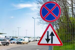 Roadside signs indicating no parking and two-way traffic, mounted on a fence near a busy parking area under a clear sky. photo