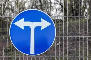 Blue circular road sign indicating a mandatory left or right turn, mounted on a wire fence with a wooded area in the background. photo