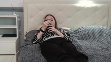 Young woman hiding behind smartphone screen while resting on bed, portraying digital generation's social media connection and privacy in bedroom environment video