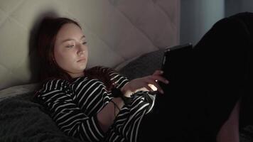 Teenage girl relaxing in bed at night, digital device casting soft screen glow across illuminated face while browsing digital content with wireless connection video