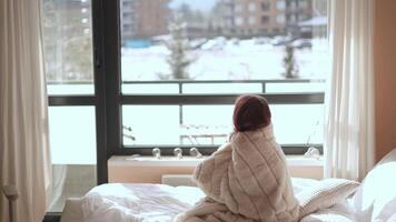 On a chilly winter morning, a woman wrapped in a cozy blanket gazes at the serene snowy landscape outside her window. She reflects on her thoughts while staying warm and enjoying the stillness video