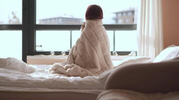 In this serene moment, an individual is captured sitting comfortably wrapped in a soft, cozy blanket, gazing thoughtfully out the window, which evokes deep feelings of tranquility and introspection video