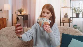 Smiling woman blogger influencer recording story on smartphone sitting on sofa showing money at home video