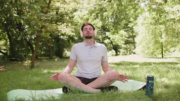 Young sporty man sitting on mat in lotus position, relaxing in city park on summer day meditation video