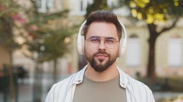 Caucasian young man taking off wireless headphones looking at camera and smiling on city street video