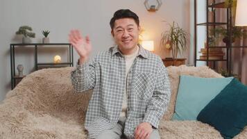 Happy young man vlogger smiling friendly at camera and waving hands while sitting on sofa at home video