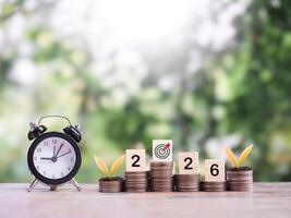 Black alarm clock, Wooden blocks with the number 2026 and plants growing up on stack of coins. The concept of saving money for manage time to success business in year 2026 photo