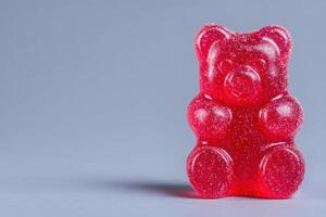 A vibrant red gummy bear displays a glossy surface and rounded features. It sits prominently on a muted gray backdrop, emphasizing its playful shape and sweet appeal. photo