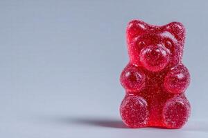 A bright red gummy bear figure, plump and textured with sugar. It sits upright on a flat surface, highlighting its cheerful expression. The neutral background enhances its vibrant color. photo
