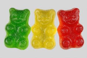 Three gummy bears in green, yellow, and red are lined up side by side. Each bear exhibits a glossy finish, showcasing their playful shapes and vibrant colors on a simple gray background. photo