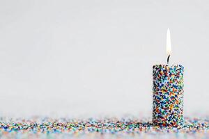 A tall, cylindrical candle adorned with colorful sprinkles stands upright on a surface covered with more sprinkles. The candle is lit, creating a warm glow in a celebratory atmosphere. photo