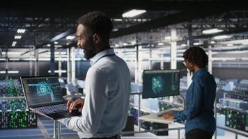 Programmer and colleague in data center reviewing AI LLM visualizations on laptop. IT coworkers analyzing neural network performance outputs to ensure stable infrastructure operations video