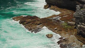 A rugged ocean shoreline where powerful waves crash against weathered rocks, creating swirling foam. Bright sunlight illuminates the stunning turquoise waters in this dynamic coastal scene. video