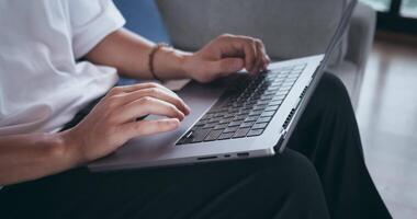 Close-up person sitting on couch at home and using laptop computer to work from home. Closeup male hands typing text on laptop, using touchpad, scrolling Internet while sitting on sofa. Work from home video