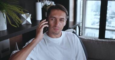 jovem homem chamando uma pessoa, ouvindo e falando em telefone com Smartphone em acolhedor sofá às casa vivo sala. relaxado homem negociando em Smartphone dispositivo. discutindo com amigo família video