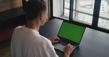 Over the shoulder shot of man's hands typing keyboard on laptop with green mock-up screen on background cozy modern office. Back view of business man using green screen laptop in office video