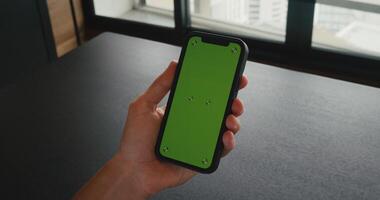 Back view of male holding chroma key green screen smartphone watching content while siting at table at office. Smartphone on hand with Green Mock-up Screen Business Concept. video