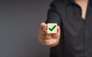 Man holds a wooden cube with a green check mark symbol while standing on a black background photo