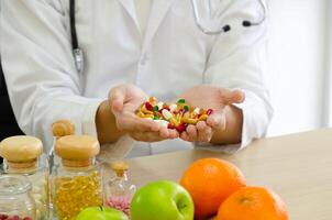 Nutritionist doctor in white coat with stethoscope holding colorful pills in open palms. Jars of pills, apple and orange on clinic desk photo