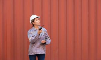 Female port worker in white helmet and uniform holding walkie talkie and tablet for coordinating logistics loading operations at container terminal shipping yard photo
