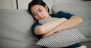 Handheld high angle view shot, A stressed young woman is leaning against a sofa, hugging a pillow tightly, her face tense with worry in a quiet room video