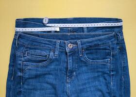 Two blue jeans of different size with measuring tape flat lay on a yellow background. Losing weight, overweight and healthy lifestyle concept photo