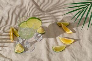 Summer day scene with refreshing drink, lime and lemon slices and tropical palm leaf shadow on sand color linen background. Sun and shadows. Minimal cocktail concept. Trendy summertime party idea. photo