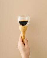 A unique conceptual photo of female hand holding an ice cream cone with cup of coffee on beige background. Minimal surreal food and drink concept. Trendy ice cream idea. Coffee aesthetic.