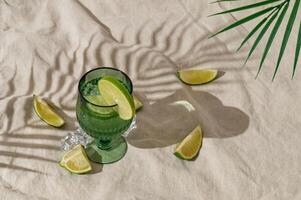 Summer day scene with exotic refreshing drink, lime slices and tropical palm leaf shadow on sand color linen background. Sun and shadows. Minimal cocktail concept. Trendy summertime party idea. photo
