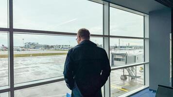 A contemplative Caucasian man gazes through rain kissed airport windows, embodying wanderlust and introspection, akin to World Tourism Day photo