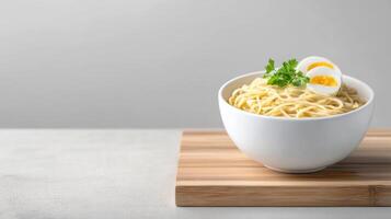 Delicious bowl of steaming hot noodle soup topped with soft boiled egg and fresh herbs served on a wooden cutting board on a plain gray background photo