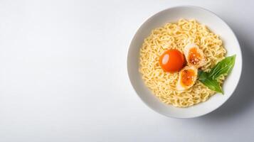 Close-up of a white bowl filled with cooked instant noodles topped with raw eggs and fresh basil leaves, on a white surface with empty space in background photo