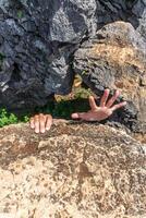 Two hands reach up from behind a rock formation, with an open palm, showing vulnerability and struggle. photo