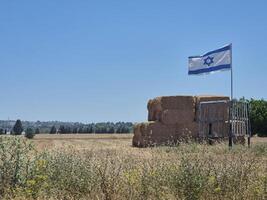 A field with hay bales stacked beside an Israeli flag, under a clear summer sky and surrounded by natural rural landscape photo