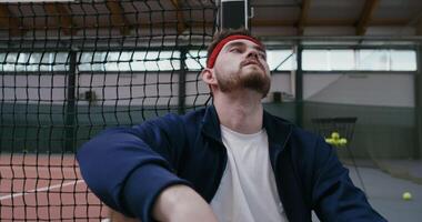 A young man is resting after training, leaning his back and head on a tennis net stretched in the middle of the court, close-up photo