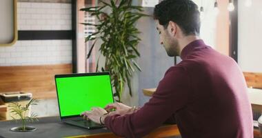 A young brunette man with stubble on his face is concentrating on typing on a laptop with a green screen, sitting at a table in a coffee shop half turned to the camera. Chroma key, copy space photo